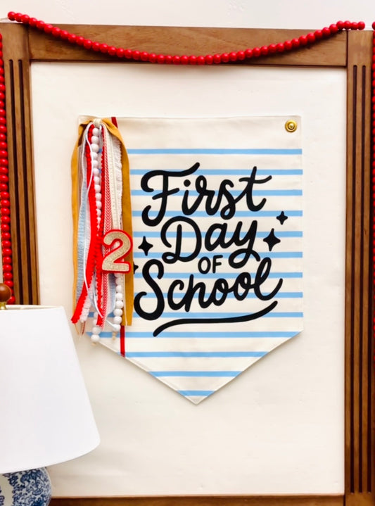 A fabric pennant banner with the text 'First Day of School' in black script over light blue and white horizontal stripes, hanging on a wooden board. The banner has a small gold grommet at the top right, a red-and-white striped ribbon with tassels on the left, and a wooden '2' decoration.