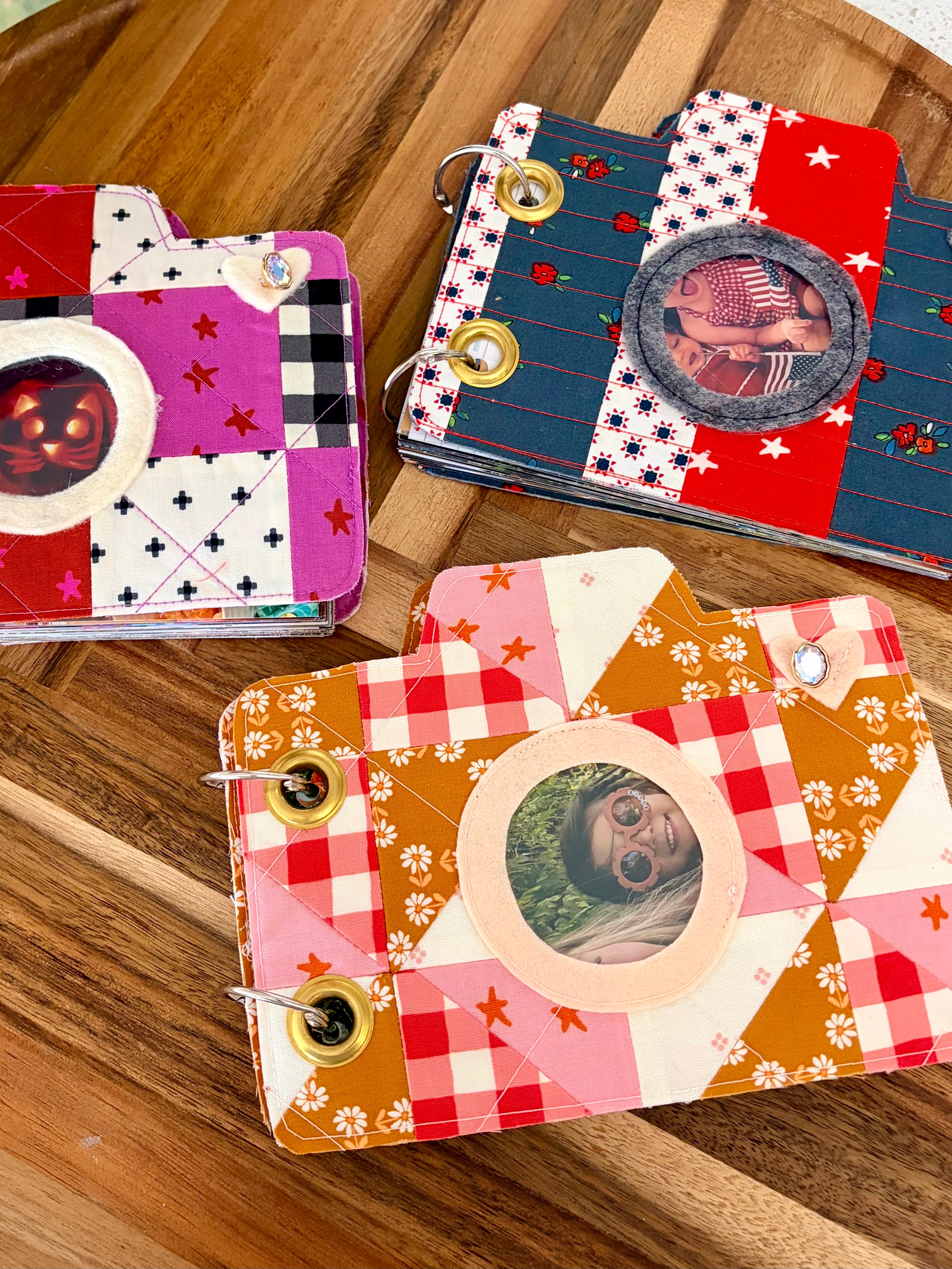 Three handmade camera-shaped ring-bound albums with colorful patchwork patterns, displayed on a wooden surface.